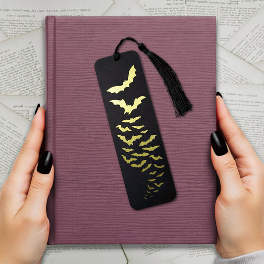 Black bookmark with a cut-out design revealing a metallic gold flock of bats in flight. Finished with a matching tassel. Perfect for gothic, spooky, and alternative readers.