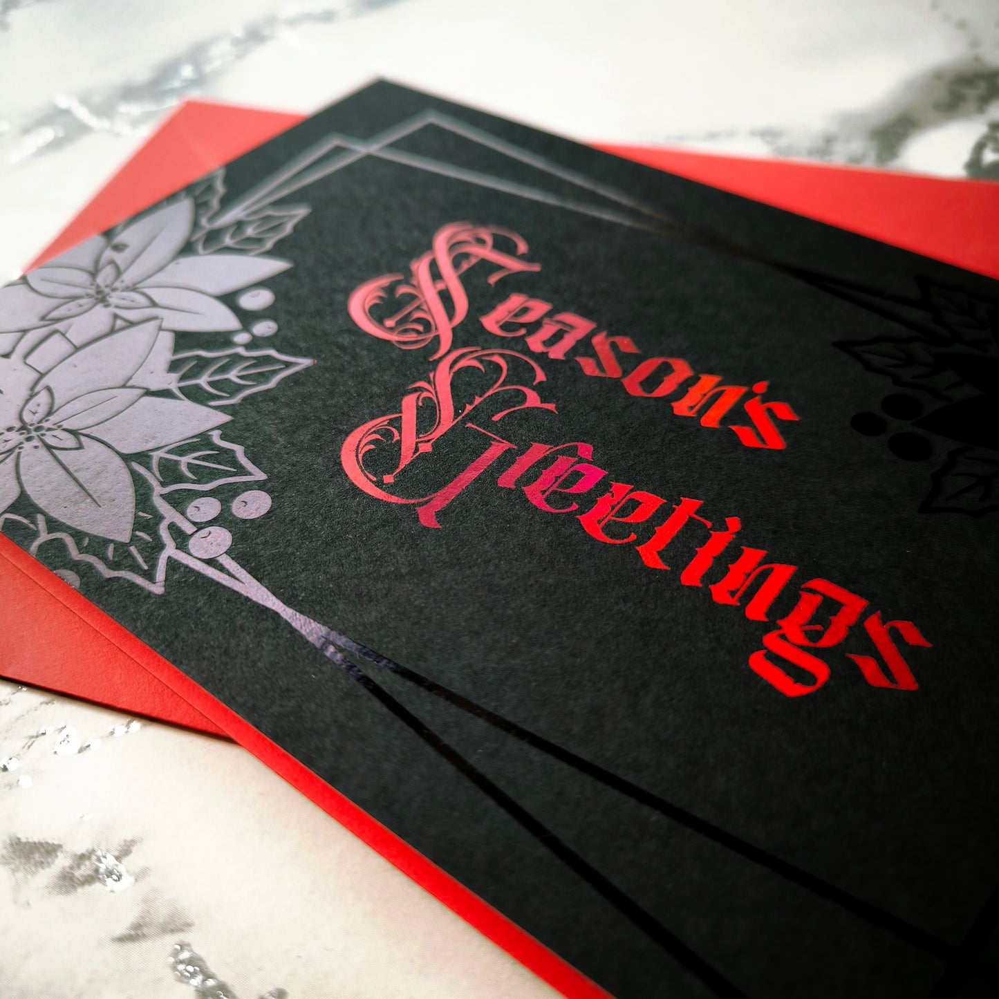 A5 landscape handmade Christmas card on black with red foil “Season’s Greetings” lettering and black foil poinsettia and holly motifs in the corners. Subtle border lines. Includes red envelope. Blank inside.