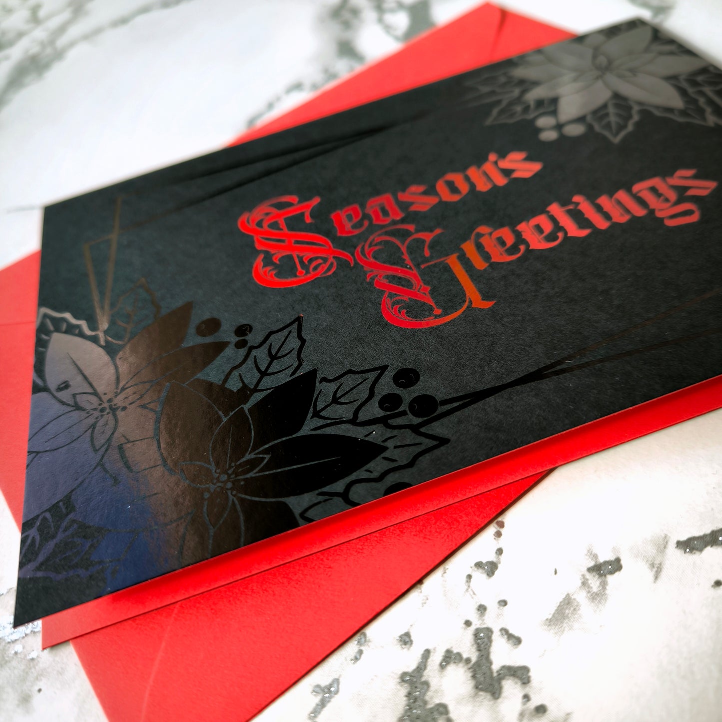 A5 landscape handmade Christmas card on black with red foil “Season’s Greetings” lettering and black foil poinsettia and holly motifs in the corners. Subtle border lines. Includes red envelope. Blank inside.