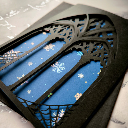 A5 handmade layered papercut Christmas card with black gothic cathedral arch window cutout and spiderweb corners, midnight blue background with holographic snowflakes. Portrait format. Includes black envelope. Blank inside.