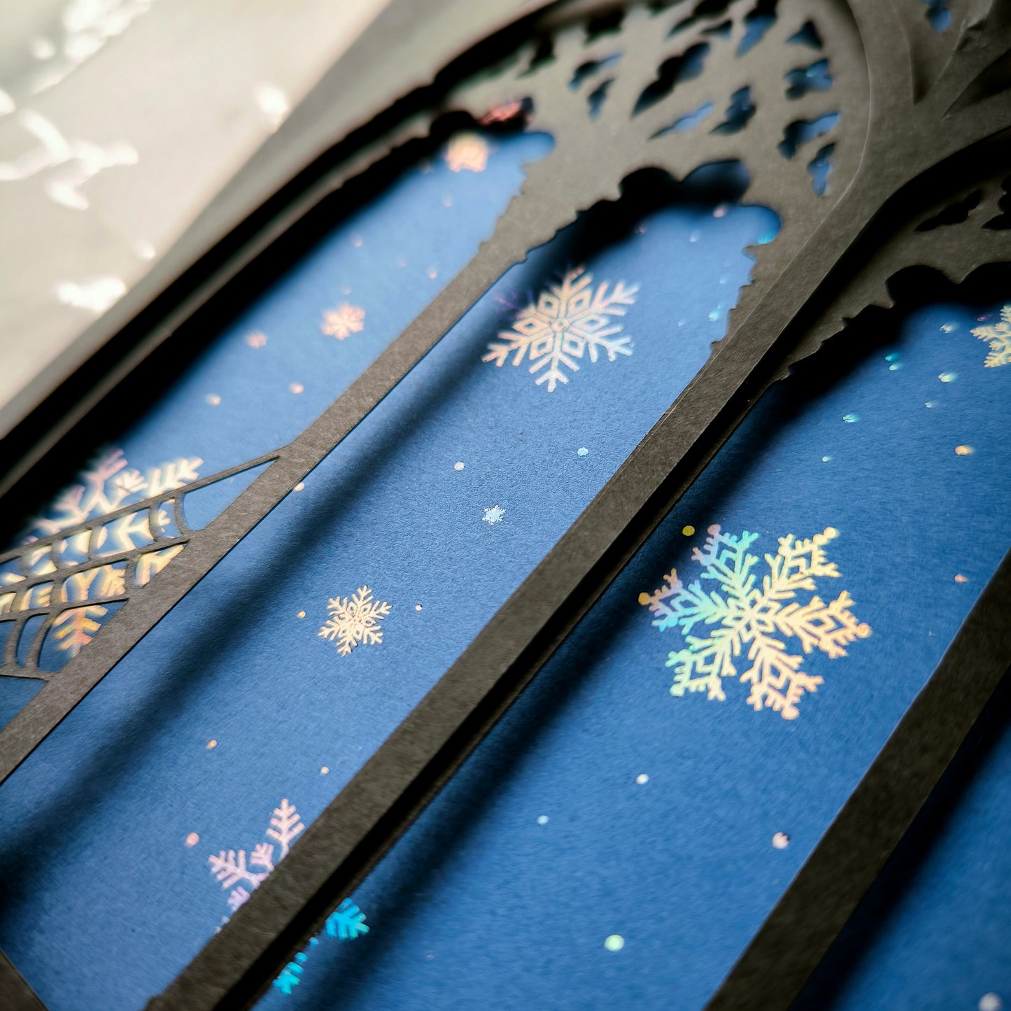 A5 handmade layered papercut Christmas card with black gothic cathedral arch window cutout and spiderweb corners, midnight blue background with holographic snowflakes. Portrait format. Includes black envelope. Blank inside.