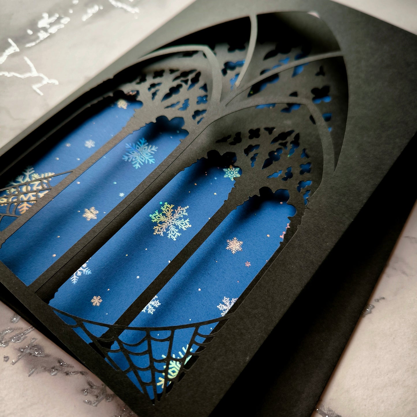 A5 handmade layered papercut Christmas card with black gothic cathedral arch window cutout and spiderweb corners, midnight blue background with holographic snowflakes. Portrait format. Includes black envelope. Blank inside.