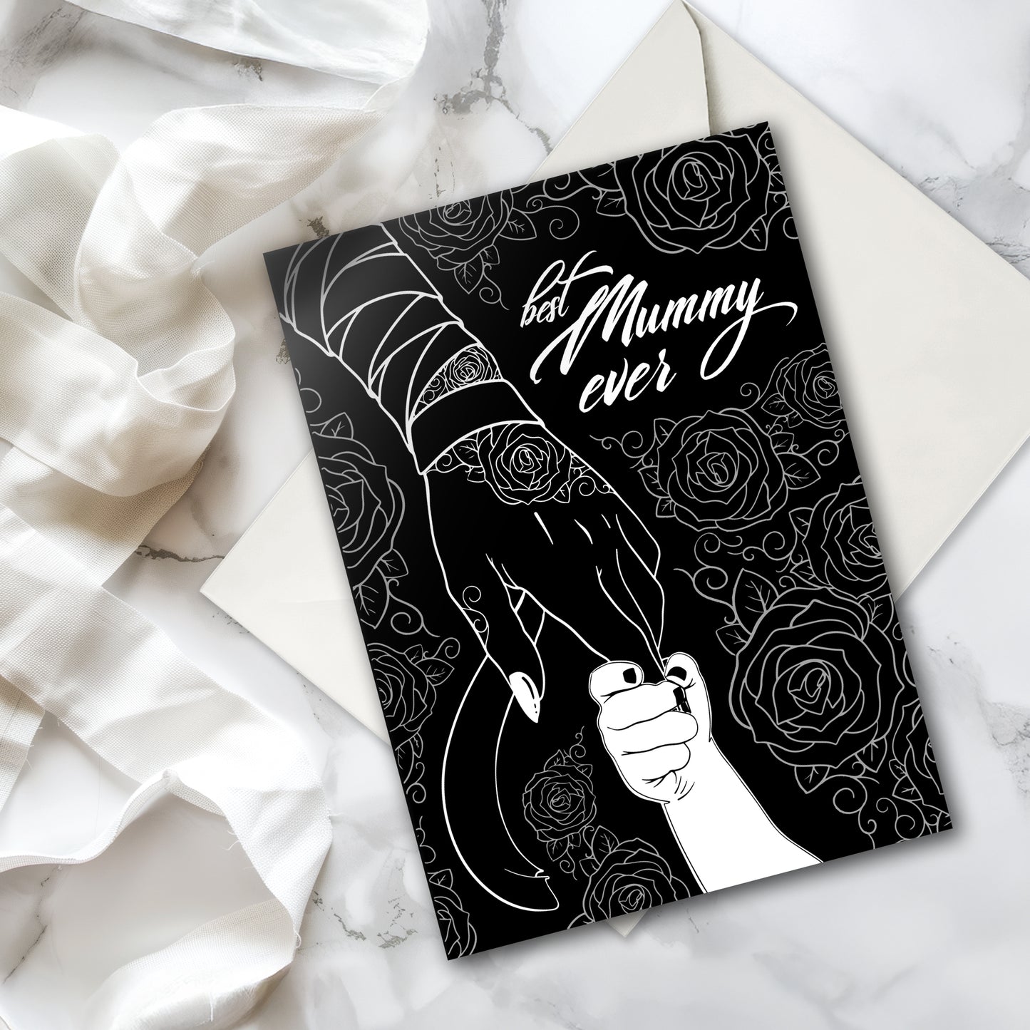 A large A5 glossy black greeting card featuring a tattoo-inspired illustration of a baby’s hand reaching for a woman’s hand wrapped in mummy bandages with rose tattoos. The card is framed by tattoo-style rose artwork. The inside is blank with matching rose motifs. Comes with an envelope included.
