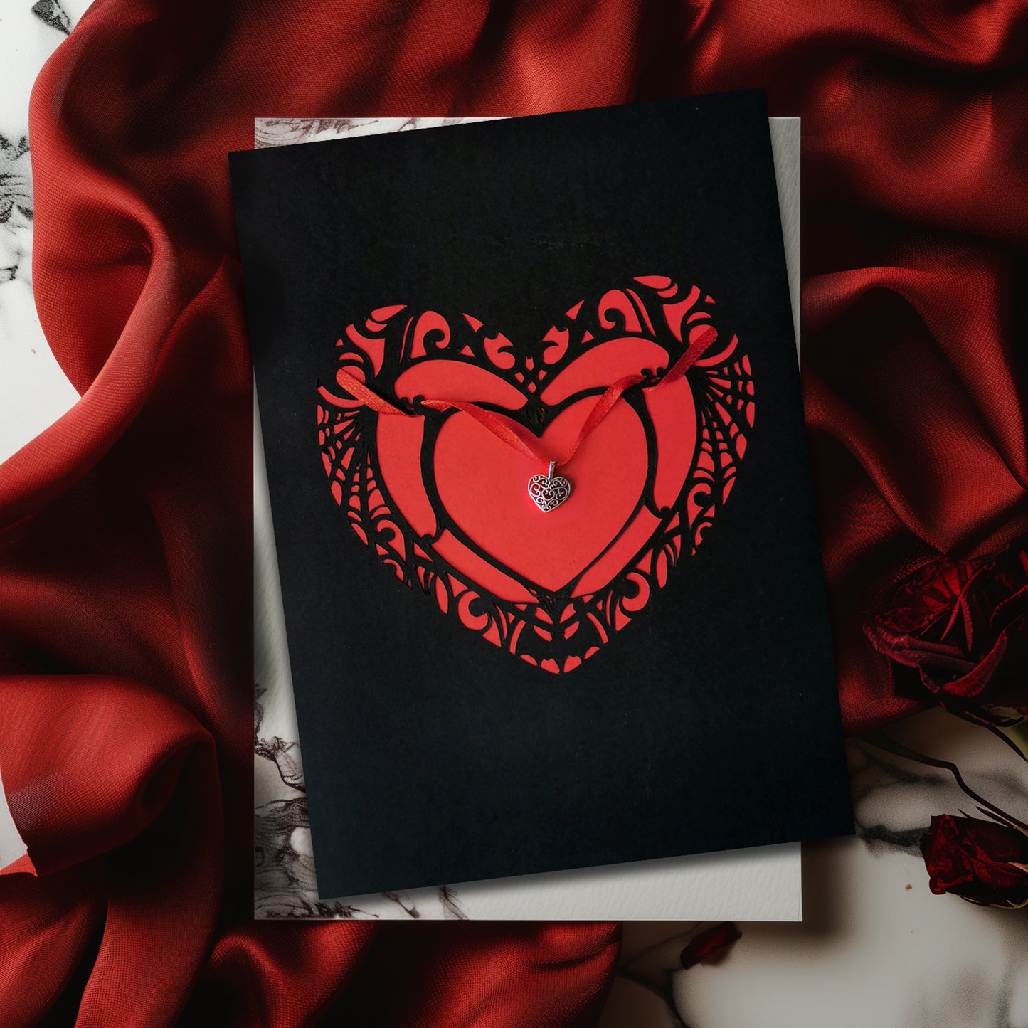 Handmade A5 black card with a red cut-out heart design and hanging silver heart charm on red ribbon. Blank white interior with envelope included. Perfect for gothic weddings, anniversaries, or alternative Valentine’s Day.