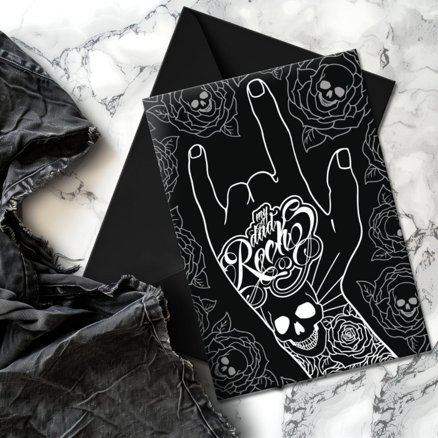 Glossy black Father’s Day card featuring a hand throwing the rock horns with skull and rose tattoos, and the phrase “My Dad Rocks” in ink lettering.