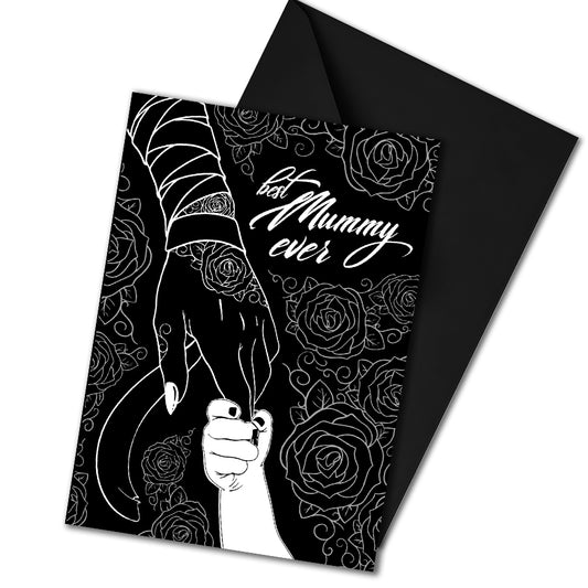 A large A5 glossy black greeting card featuring a tattoo-inspired illustration of a baby’s hand reaching for a woman’s hand wrapped in mummy bandages with rose tattoos. The card is framed by tattoo-style rose artwork. The inside is blank with matching rose motifs. Comes with an envelope included.
