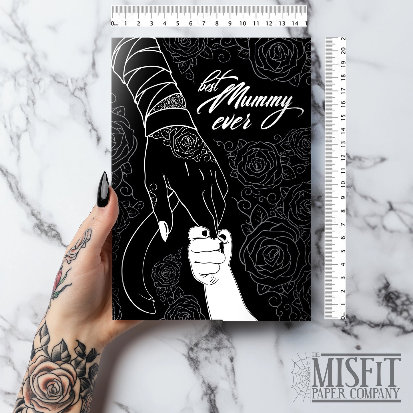 A large A5 glossy black greeting card featuring a tattoo-inspired illustration of a baby’s hand reaching for a woman’s hand wrapped in mummy bandages with rose tattoos. The card is framed by tattoo-style rose artwork. The inside is blank with matching rose motifs. Comes with an envelope included.