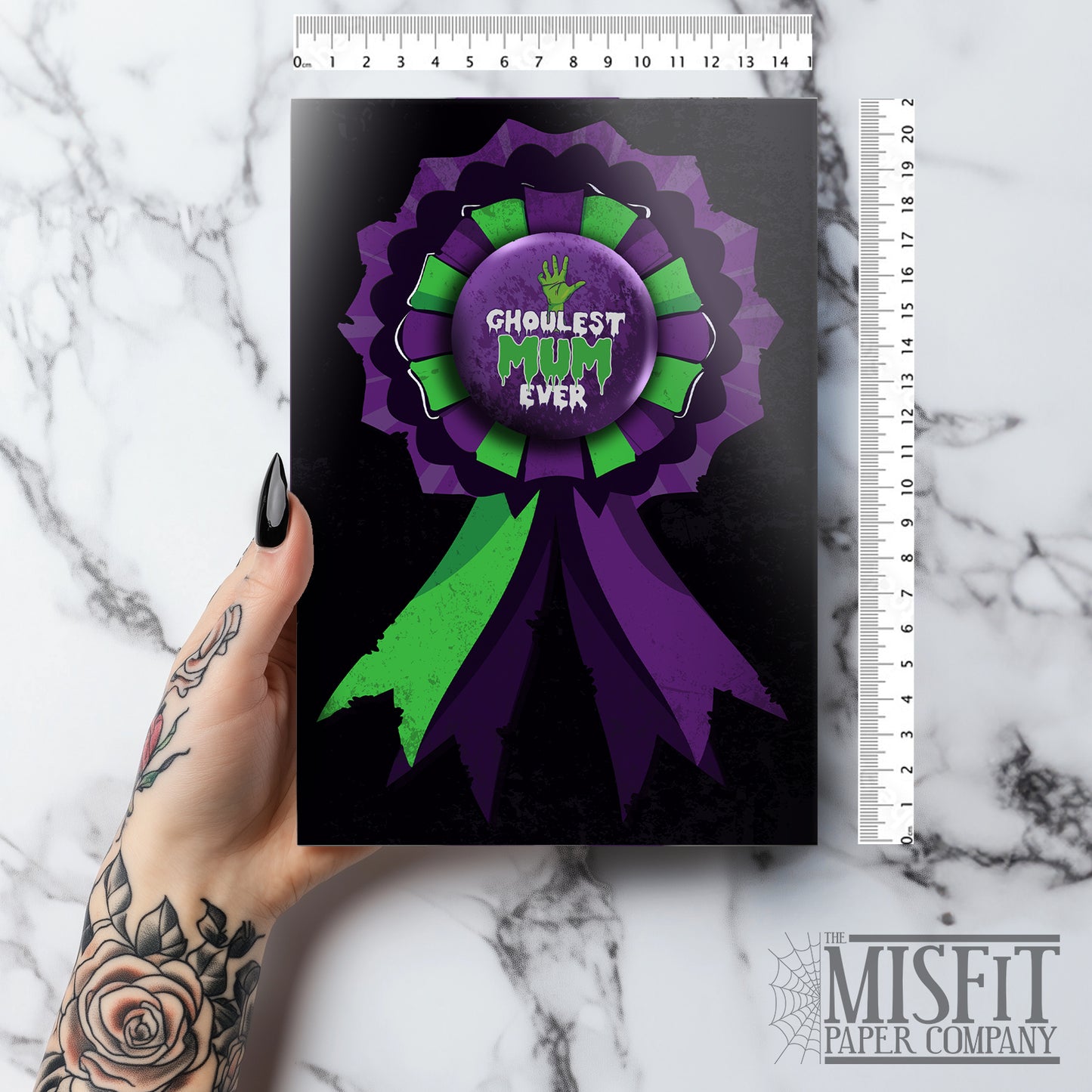 A large glossy A5 greeting card featuring a green and purple rosette with a removable 58mm badge reading "Ghoulest Mom Ever" in a bold, spooky font. Available in Mom, Mum, or Mam variations. The card is blank inside and comes with an envelope.