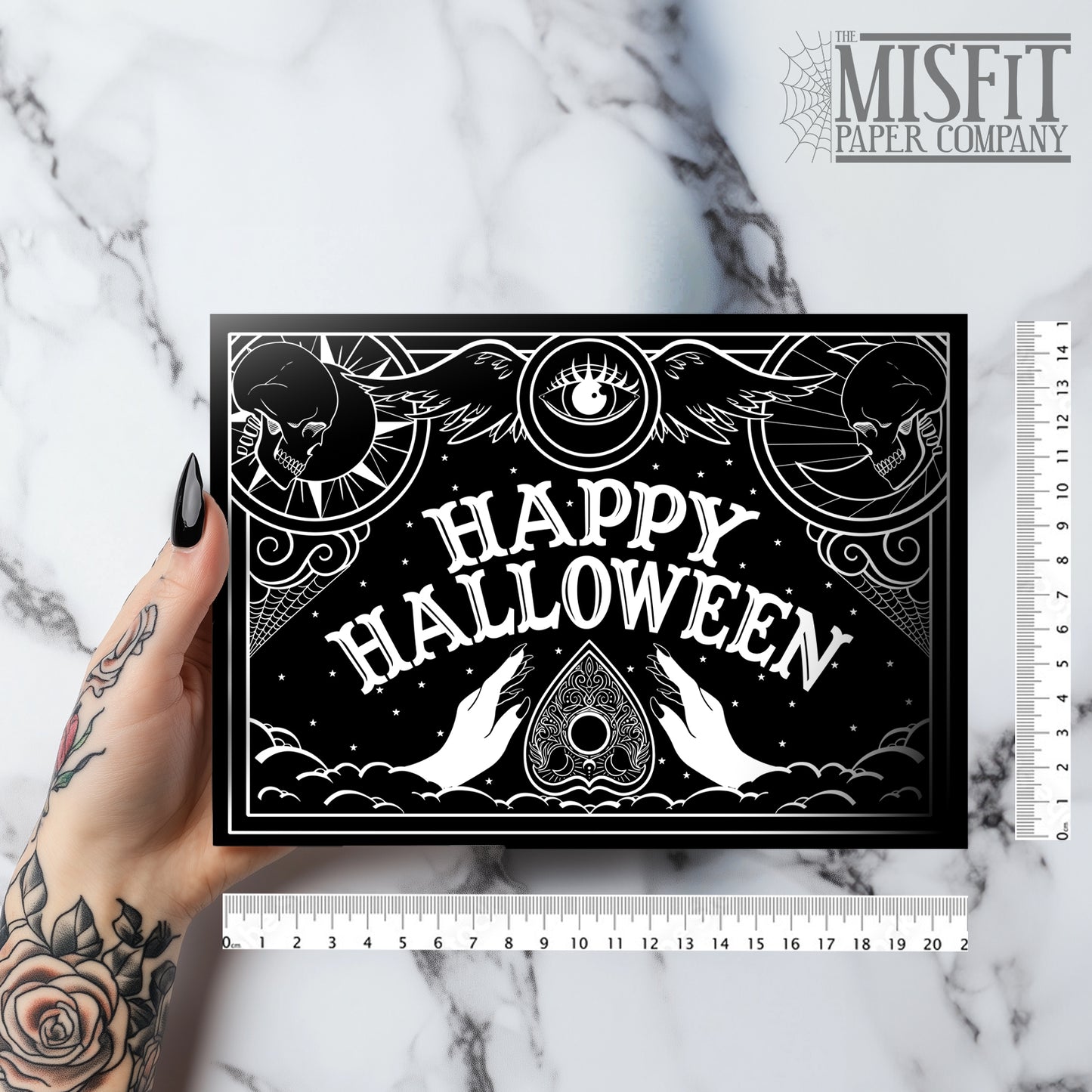 A black glossy A5 Halloween card with a white Ouija board-inspired “Happy Halloween” design, styled with gothic props on a dark background.