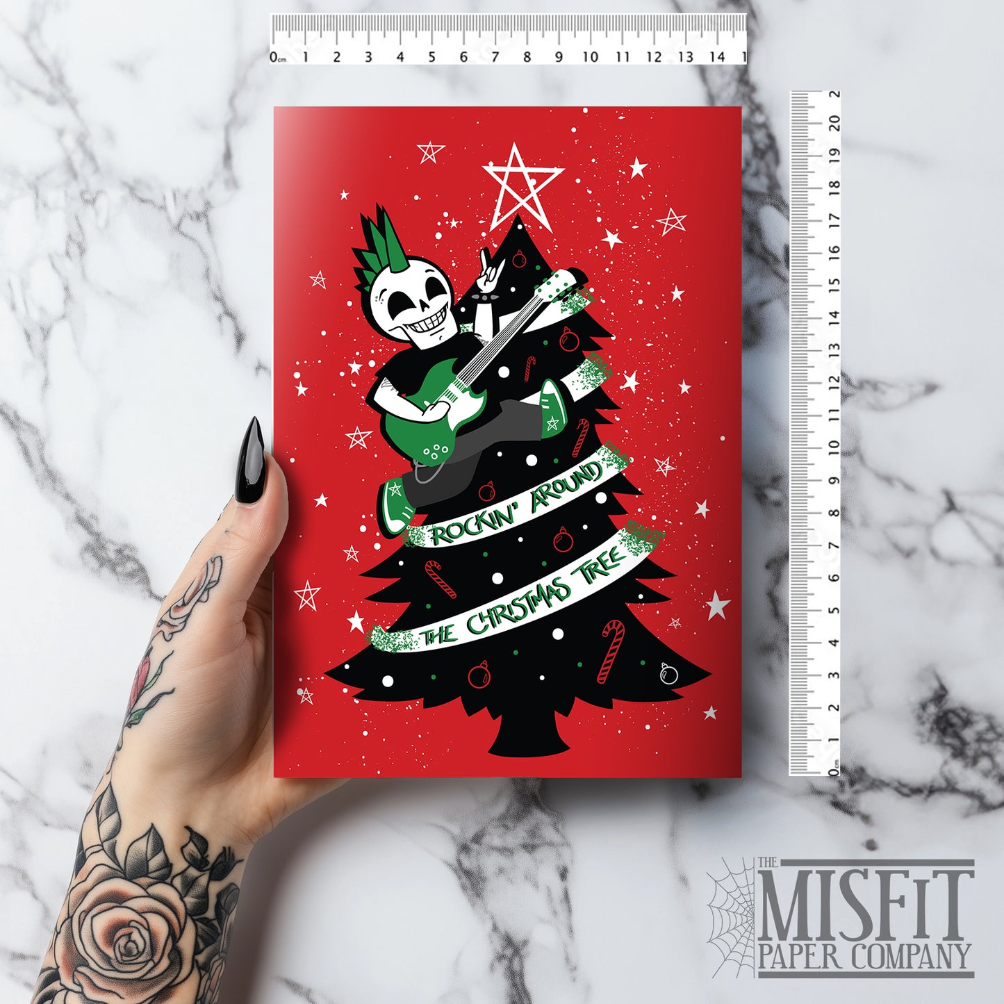A5 glossy portrait Christmas card on red with black tree, white star and green ribbon reading “Rockin’ around the Christmas tree”. Skeleton with green mohawk plays electric guitar. Includes red envelope. Inside has small red star print with blank area.