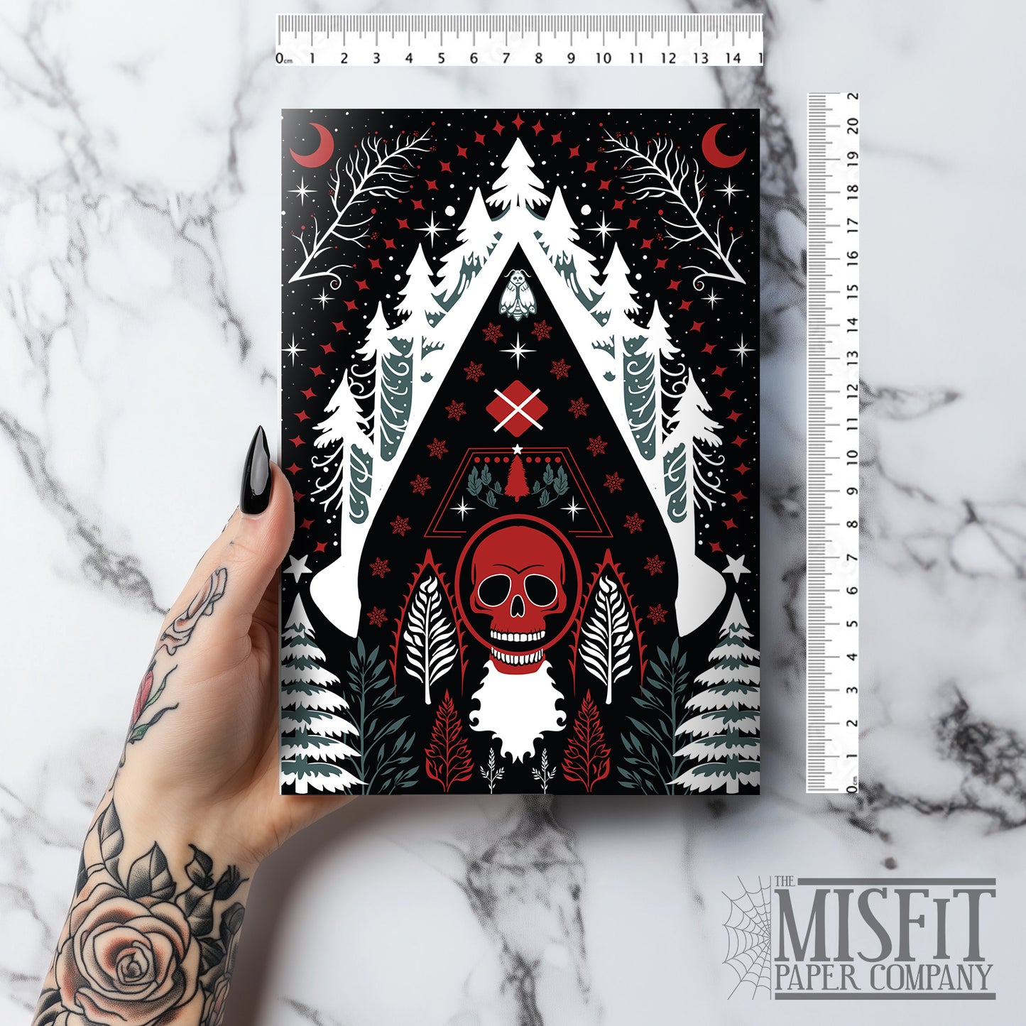 A5 glossy portrait greeting card with Scandi folk art design. Black background, white snowy pines, red crescents, stars, and a central red skull Santa motif. Graphic botanical details. Includes red envelope. Blank inside.