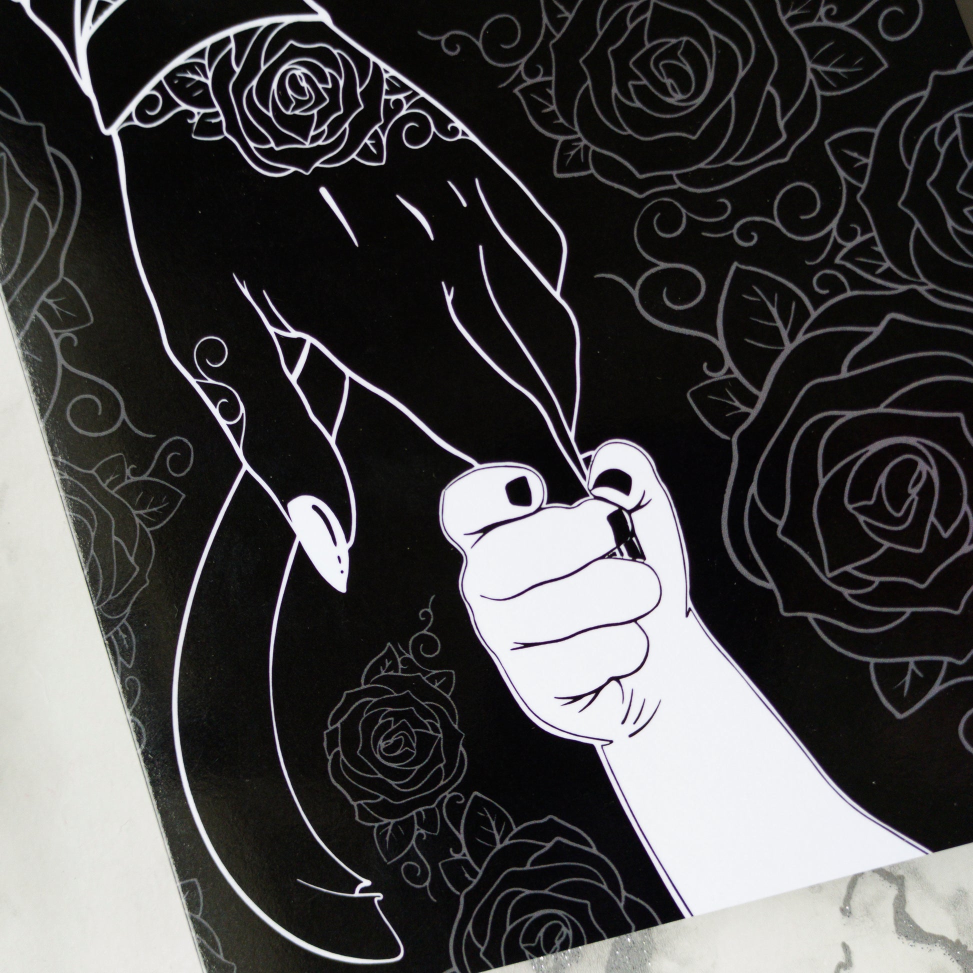 A large A5 glossy black greeting card featuring a tattoo-inspired illustration of a baby’s hand reaching for a woman’s hand wrapped in mummy bandages with rose tattoos. The card is framed by tattoo-style rose artwork. The inside is blank with matching rose motifs. Comes with an envelope included.