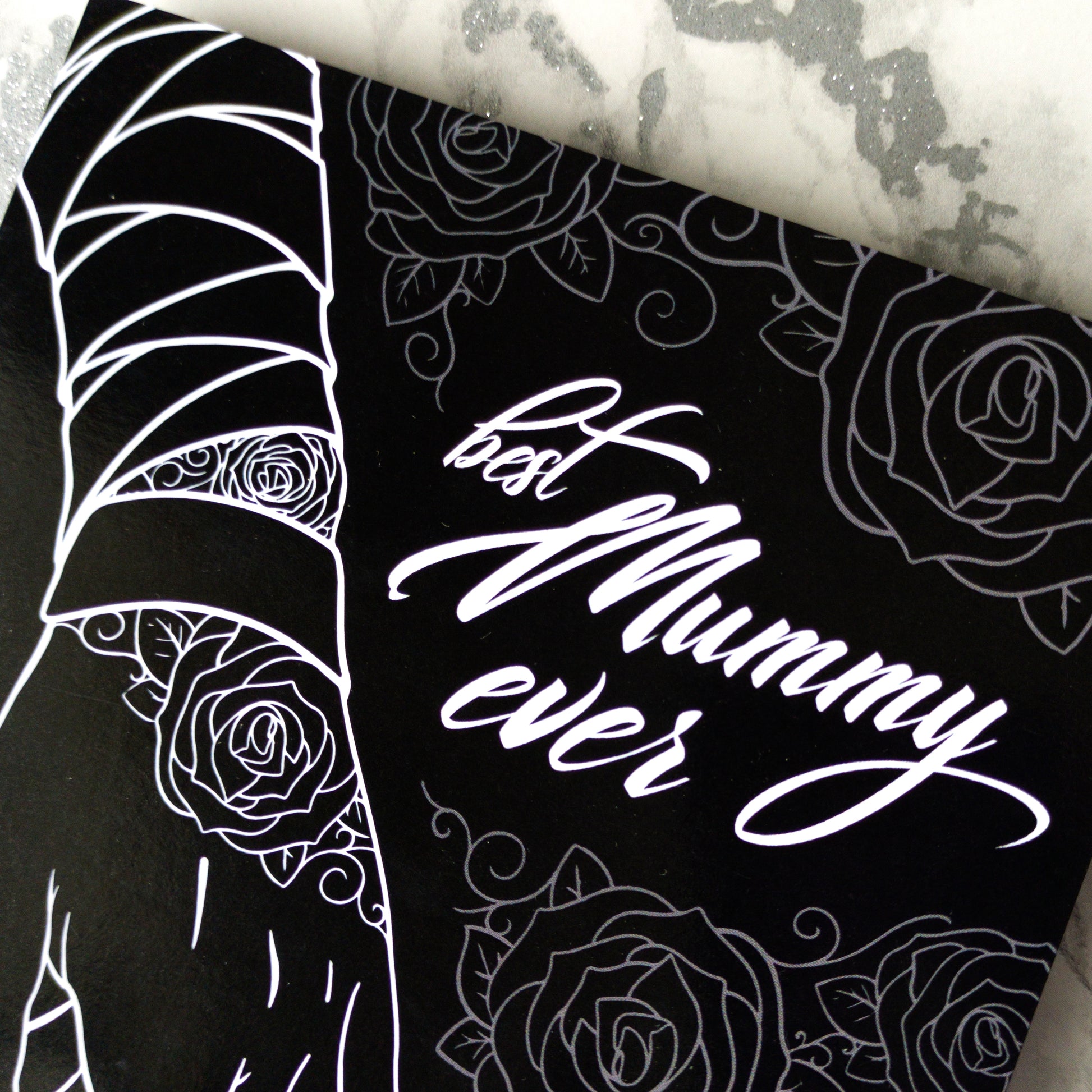 A large A5 glossy black greeting card featuring a tattoo-inspired illustration of a baby’s hand reaching for a woman’s hand wrapped in mummy bandages with rose tattoos. The card is framed by tattoo-style rose artwork. The inside is blank with matching rose motifs. Comes with an envelope included.