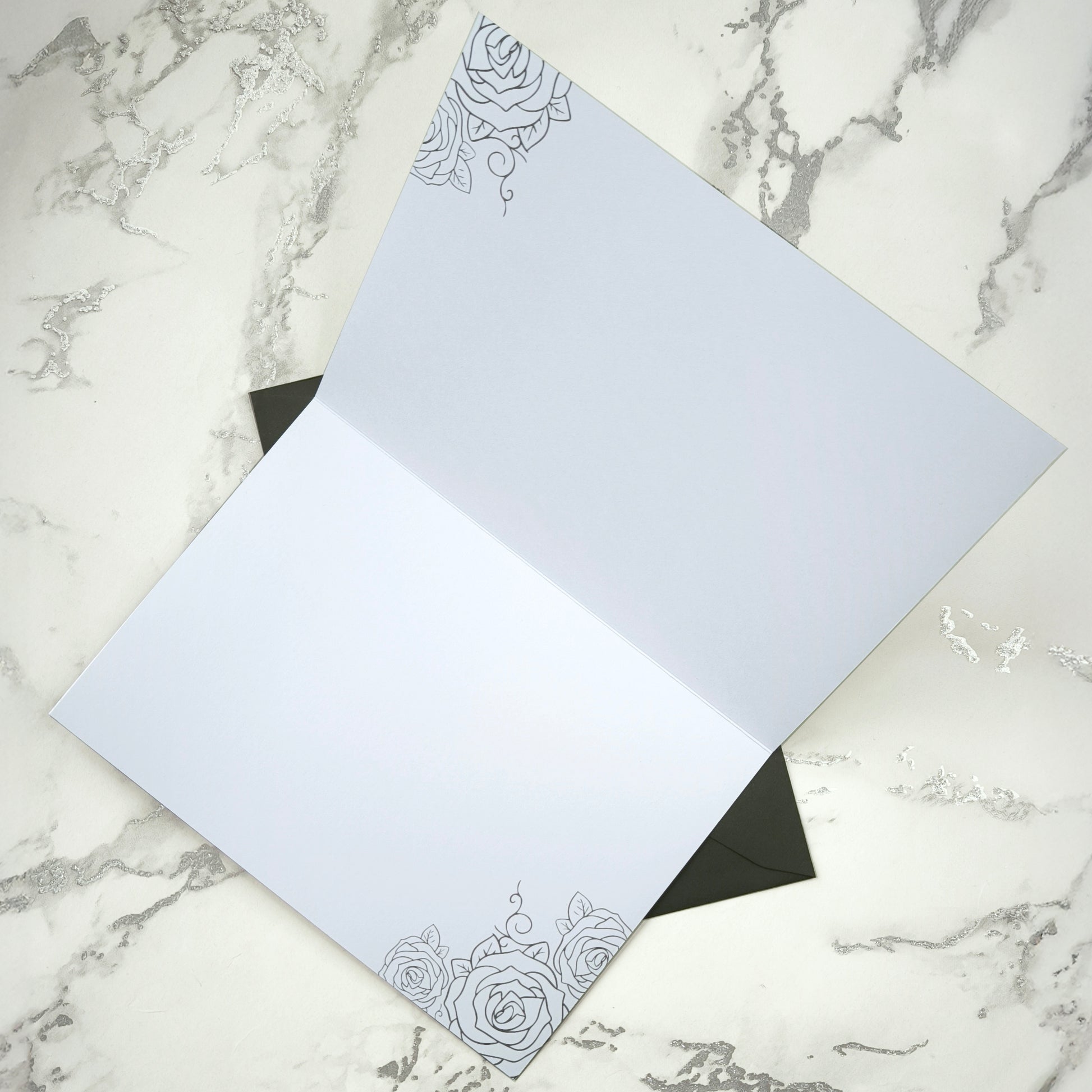 A large A5 glossy black greeting card featuring a tattoo-inspired illustration of a baby’s hand reaching for a woman’s hand wrapped in mummy bandages with rose tattoos. The card is framed by tattoo-style rose artwork. The inside is blank with matching rose motifs. Comes with an envelope included.