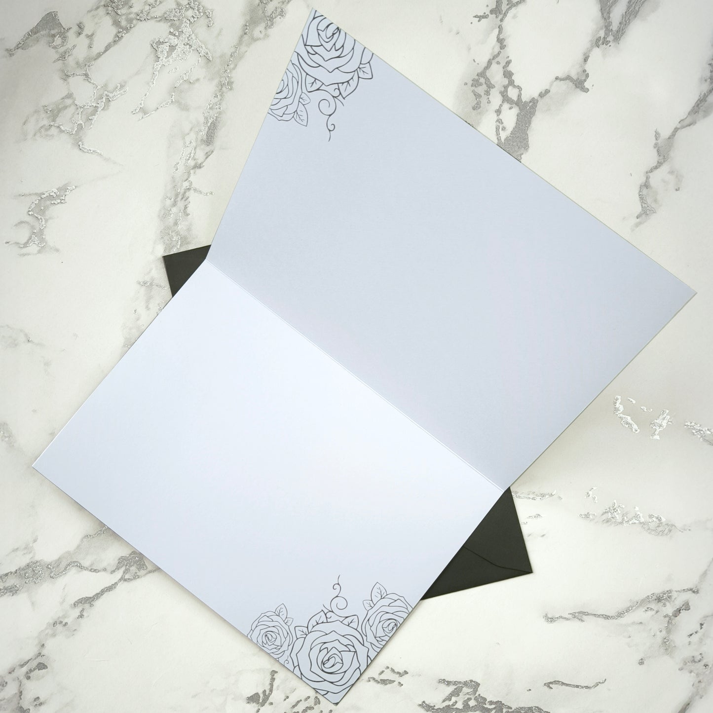 A large A5 glossy black greeting card featuring a tattoo-inspired illustration of a baby’s hand reaching for a woman’s hand wrapped in mummy bandages with rose tattoos. The card is framed by tattoo-style rose artwork. The inside is blank with matching rose motifs. Comes with an envelope included.