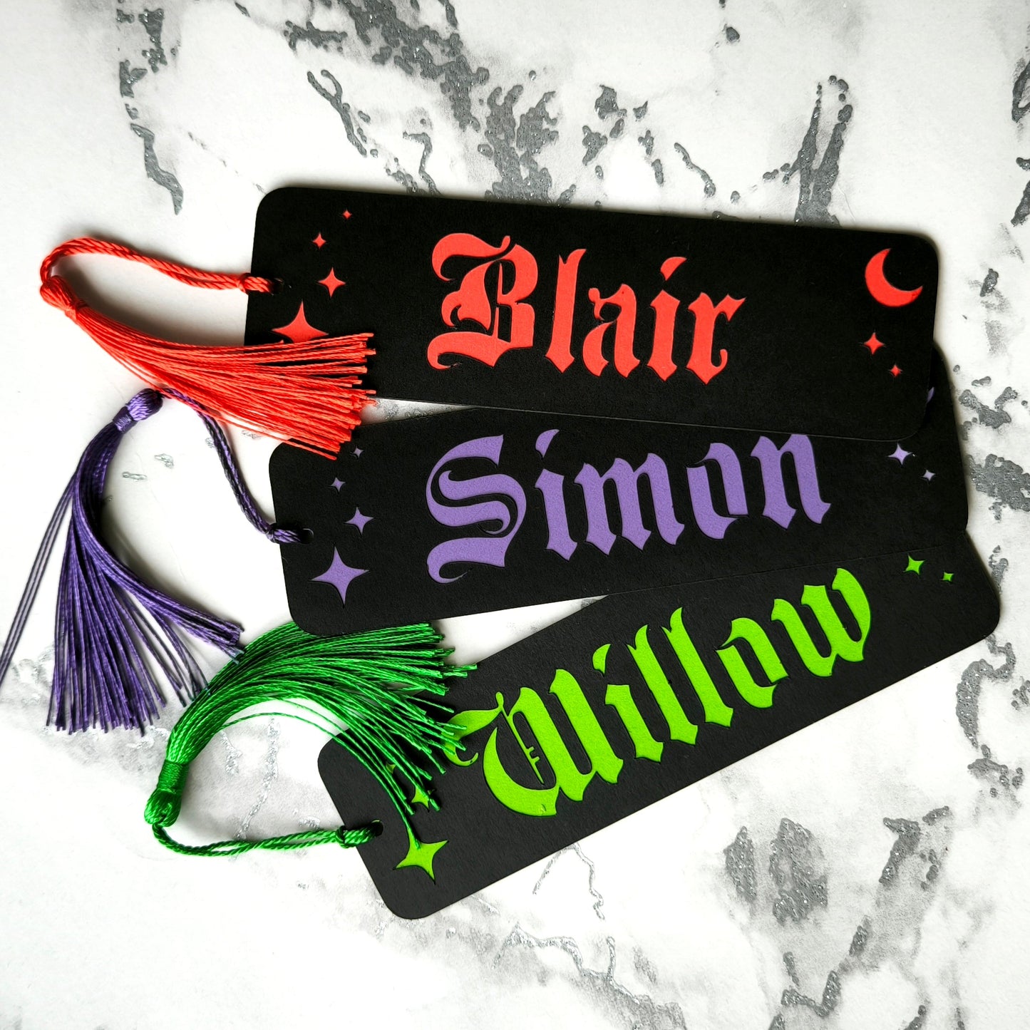 Custom gothic bookmarks with cutout names and celestial moon and star designs in red, purple, green, gold, and silver, with matching tassels