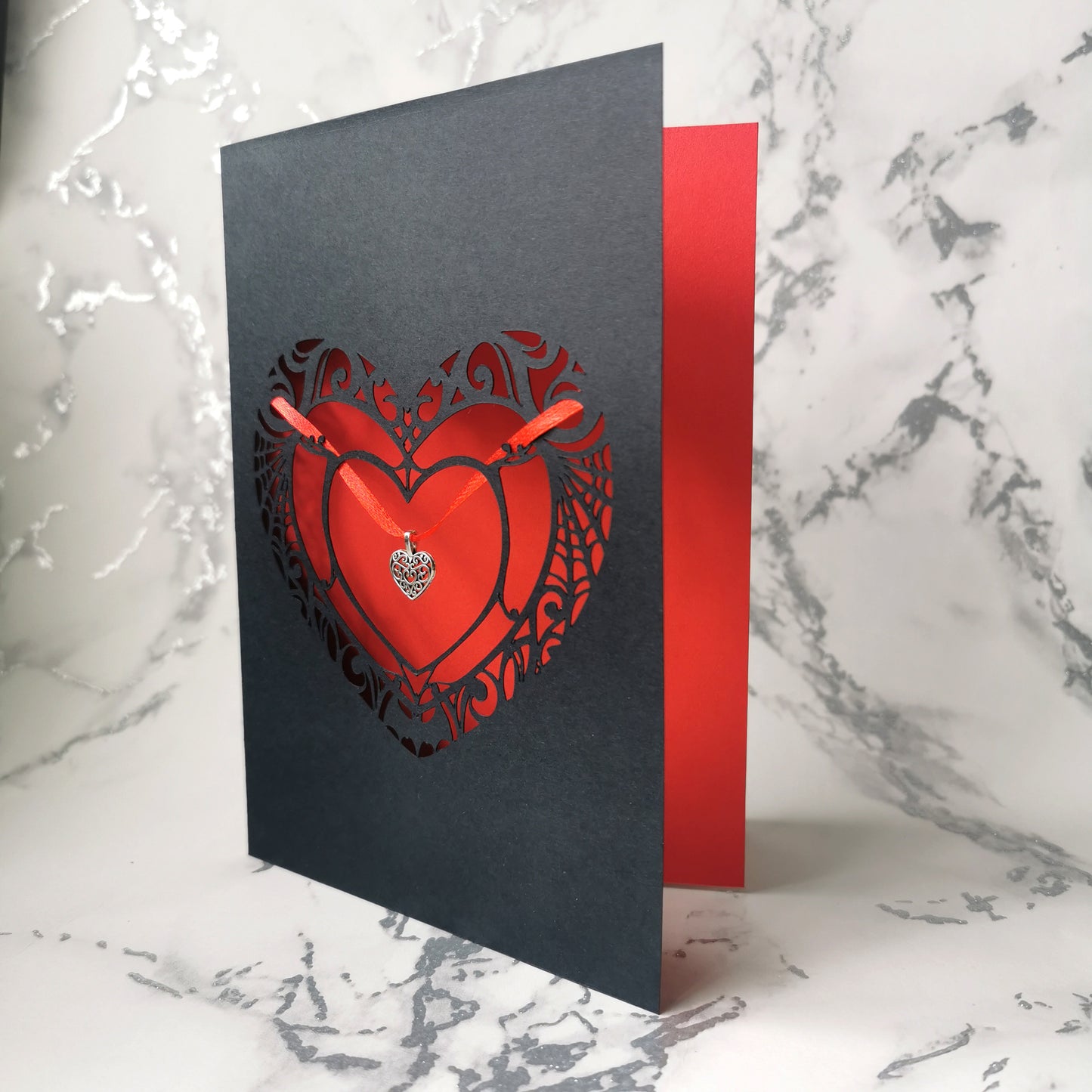 Handmade A5 black card with a red cut-out heart design and hanging silver heart charm on red ribbon. Blank white interior with envelope included. Perfect for gothic weddings, anniversaries, or alternative Valentine’s Day.