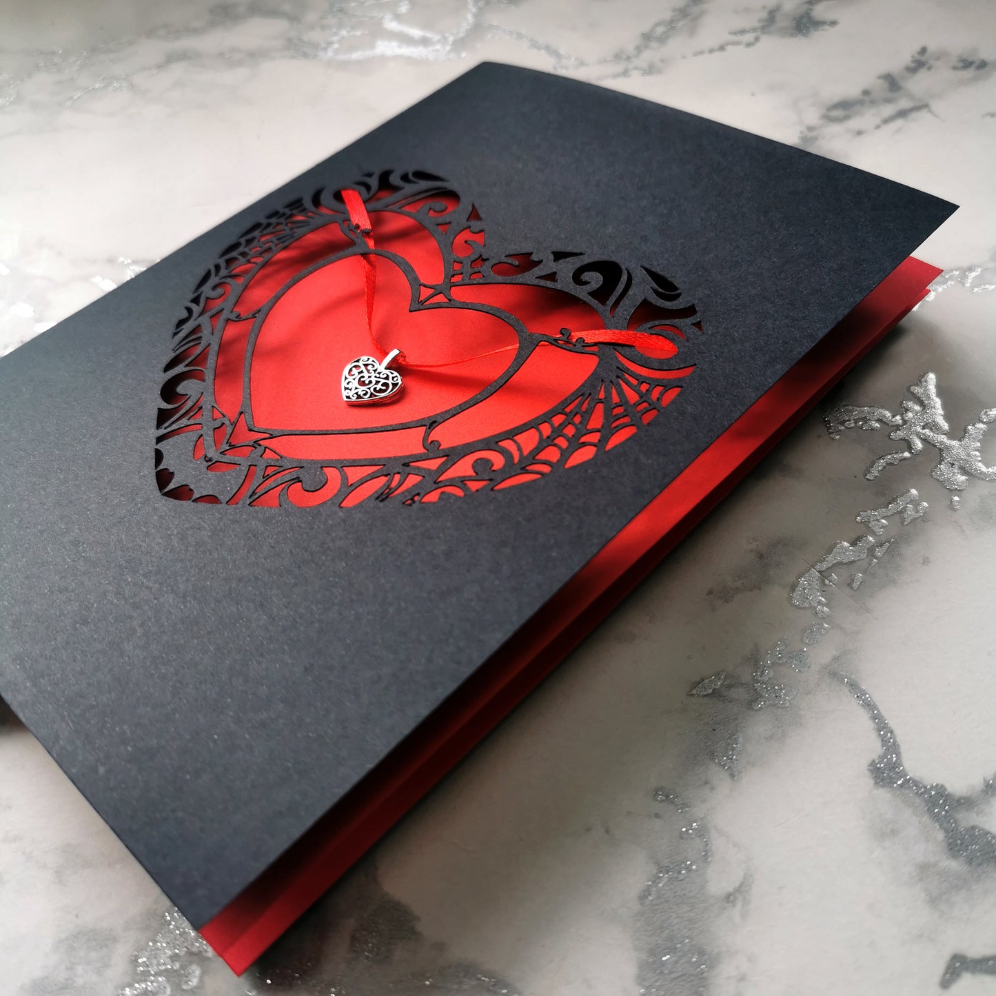 Handmade A5 black card with a red cut-out heart design and hanging silver heart charm on red ribbon. Blank white interior with envelope included. Perfect for gothic weddings, anniversaries, or alternative Valentine’s Day.