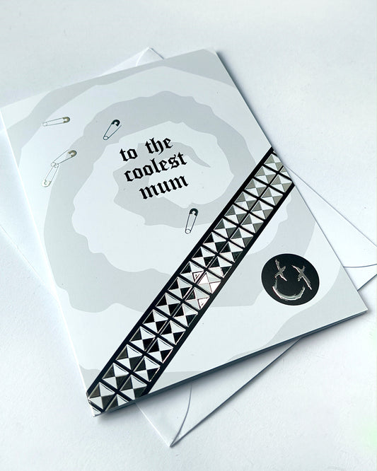 A5 greeting card reading “to the coolest mum” with gothic emo styling, featuring chrome stud belt graphic, safety pin details and a black smiley badge, paired with a white envelope.