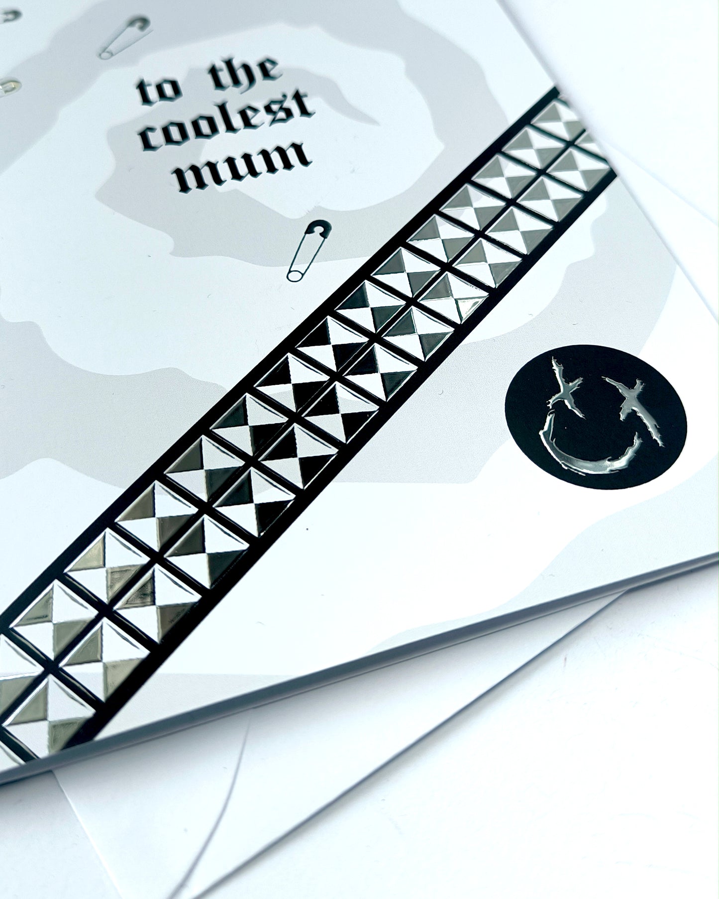 A5 greeting card reading “to the coolest mum” with gothic emo styling, featuring chrome stud belt graphic, safety pin details and a black smiley badge, paired with a white envelope.