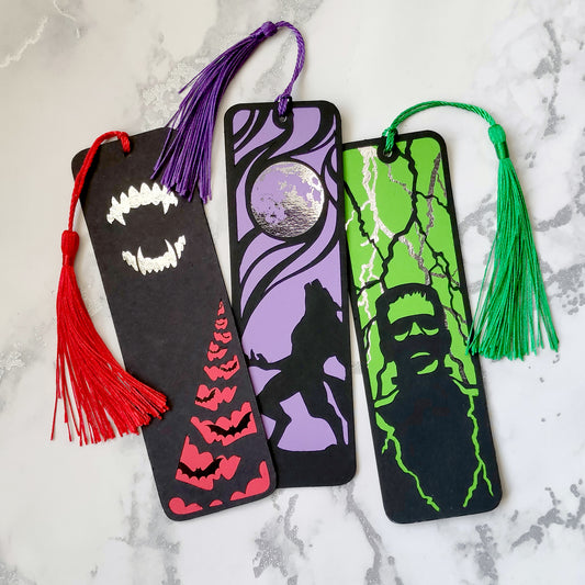 Set of 3 gothic bookmarks featuring classic horror icons: a howling werewolf, Dracula fangs, and Frankenstein’s monster. Each with dark backgrounds, metallic details, bold designs, and matching tassels. Perfect for horror fans and alternative book lovers.