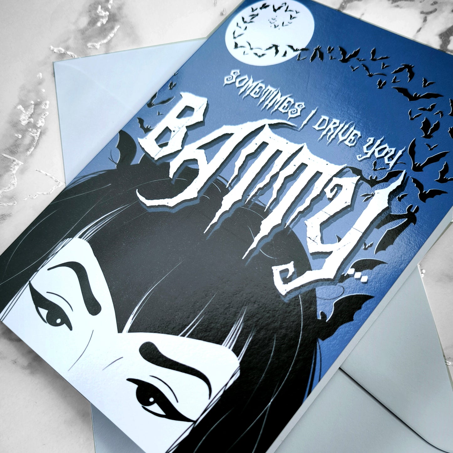 A large glossy A5 greeting card designed in a vintage horror poster style. The cover features a tired mum with bats flying over her head and the phrase "Sometimes I drive you batty...". Inside reads "But I’m your favourite vampire brat", with UK or US spelling options. Comes with a white envelope.