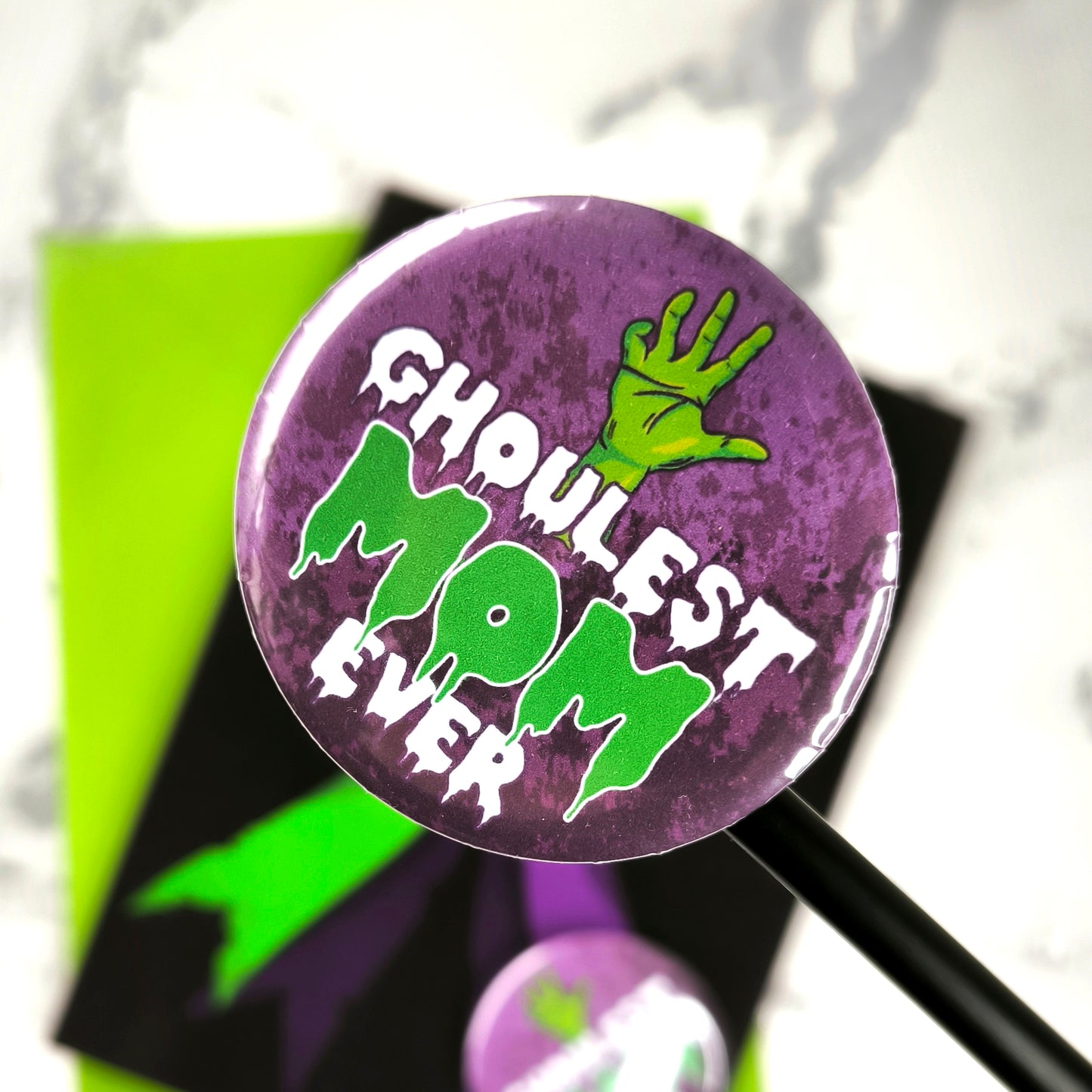 A large glossy A5 greeting card featuring a green and purple rosette with a removable 58mm badge reading "Ghoulest Mom Ever" in a bold, spooky font. Available in Mom, Mum, or Mam variations. The card is blank inside and comes with an envelope.