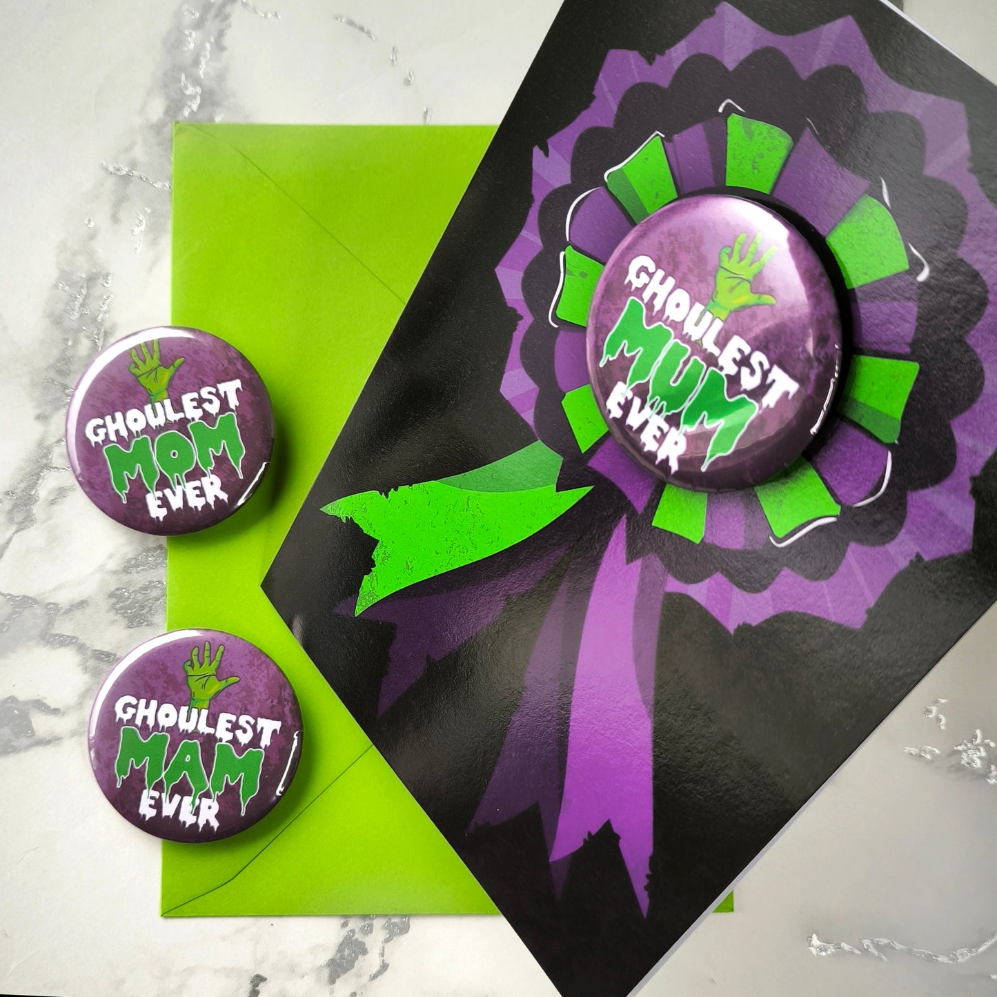 A large glossy A5 greeting card featuring a green and purple rosette with a removable 58mm badge reading "Ghoulest Mom Ever" in a bold, spooky font. Available in Mom, Mum, or Mam variations. The card is blank inside and comes with an envelope.