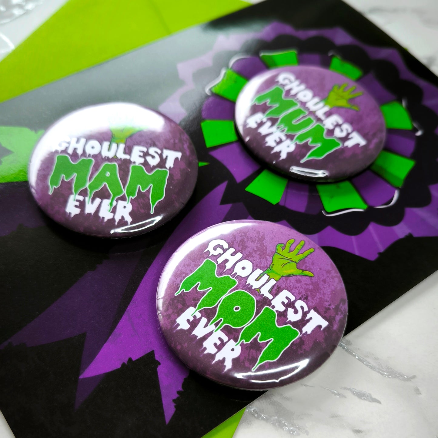 A large glossy A5 greeting card featuring a green and purple rosette with a removable 58mm badge reading "Ghoulest Mom Ever" in a bold, spooky font. Available in Mom, Mum, or Mam variations. The card is blank inside and comes with an envelope.