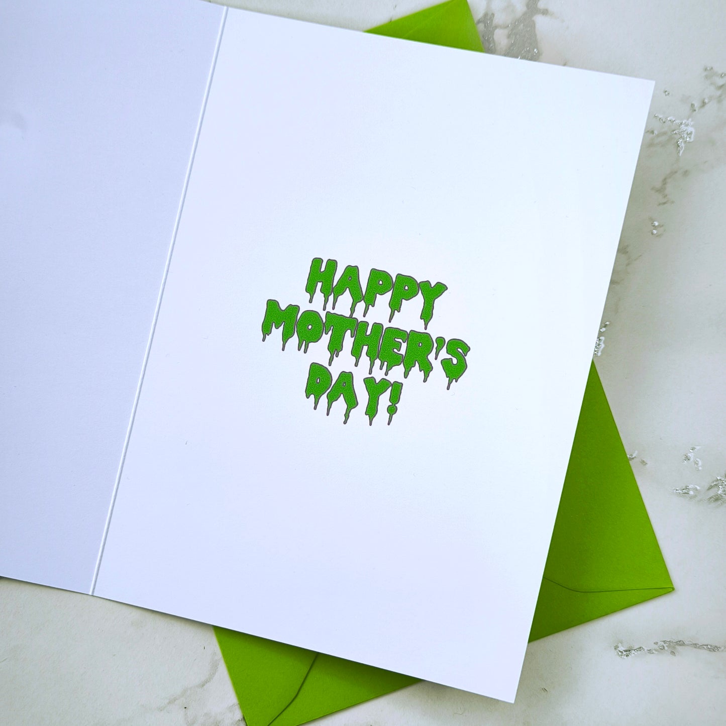 A large glossy A5 greeting card featuring a green and purple rosette with a removable 58mm badge reading "Ghoulest Mom Ever" in a bold, spooky font. Available in Mom, Mum, or Mam variations. The card is blank inside and comes with an envelope.