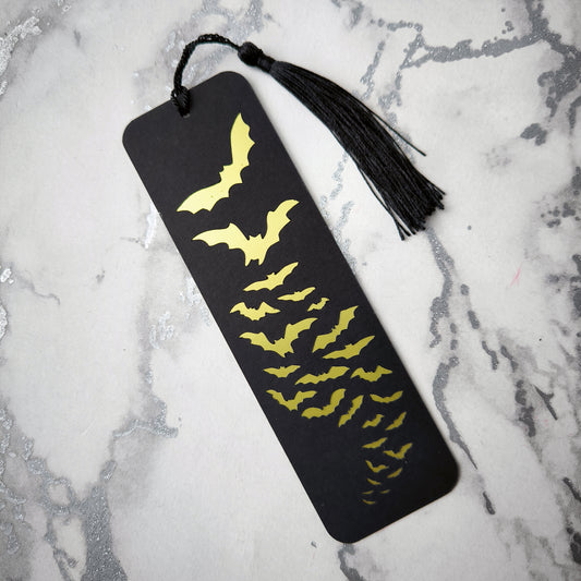 Black bookmark with a cut-out design revealing a metallic gold flock of bats in flight. Finished with a matching tassel. Perfect for gothic, spooky, and alternative readers.