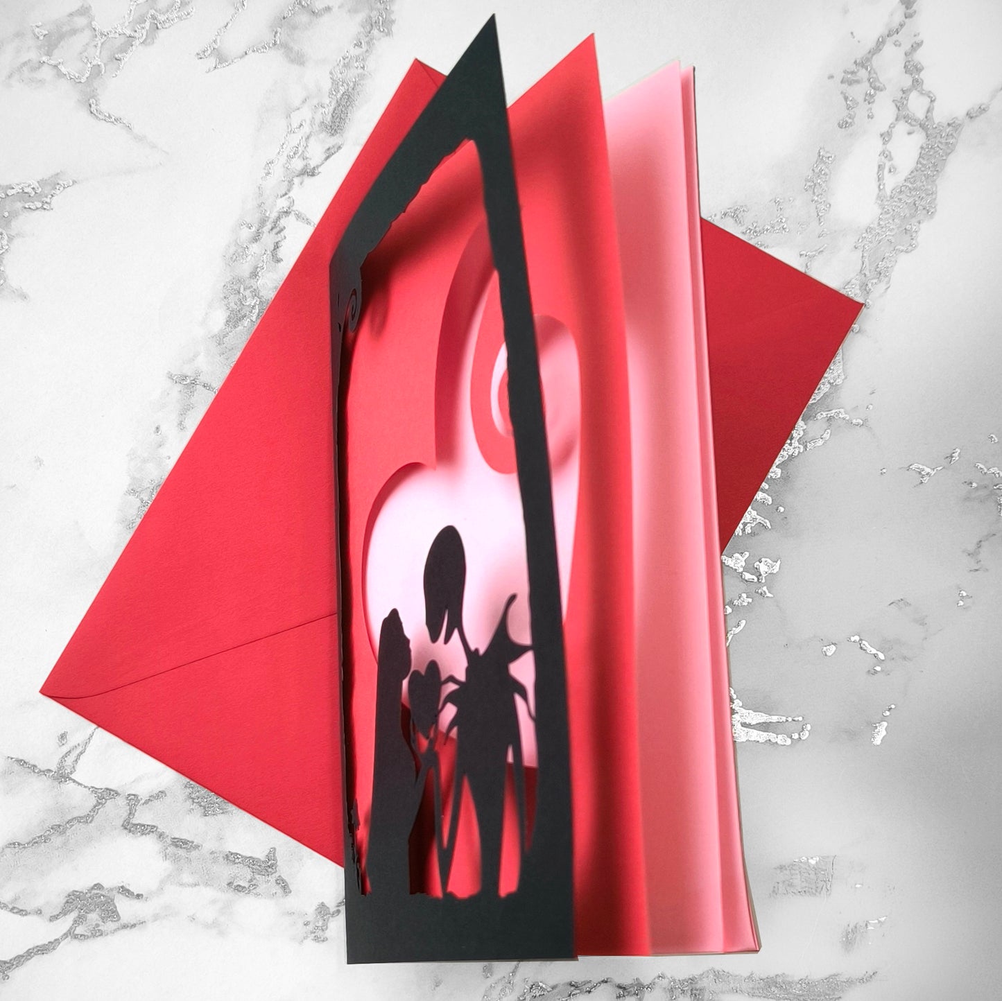 Handmade gothic romance card with black cut-out silhouette of Jack and Sally exchanging a heart, layered over a red heart with pink interior, A5 size.