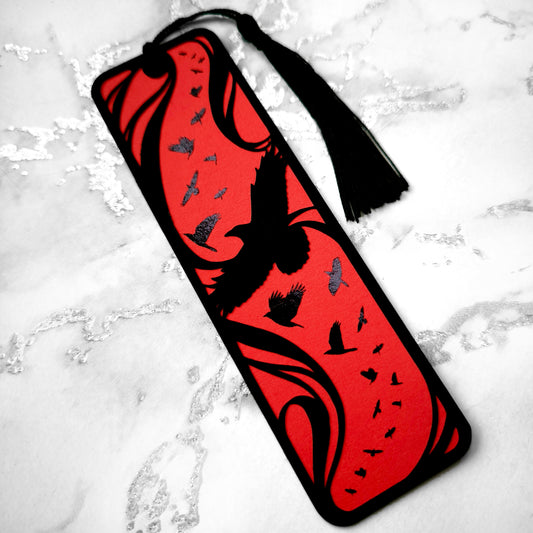 A handmade card bookmark. the black top layer is cut to a sweeping crow silhouette framed by flowing lines, set against a blood-red base scattered with glossy black-foiled crows in flight. Finished with a sleek black tassel.