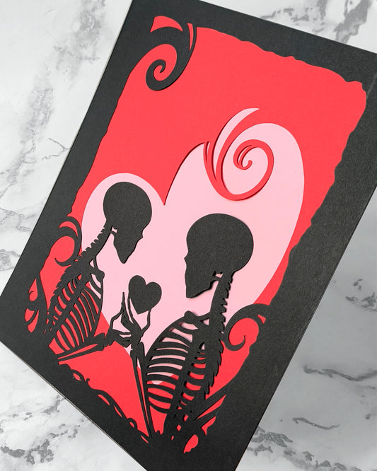 Layered papercut artwork showing two skeleton silhouettes facing each other exchanging a small heart, set in black against red and pink heart layers with gothic flourishes. measures 8 x 10 inches. frame not included.