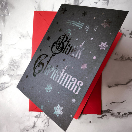 A5 handmade black Christmas card with black foil snowflakes and gothic lettering reading “Dreaming of a Black Christmas”. Portrait format on smooth black card with red interior. Includes red envelope. Blank inside.