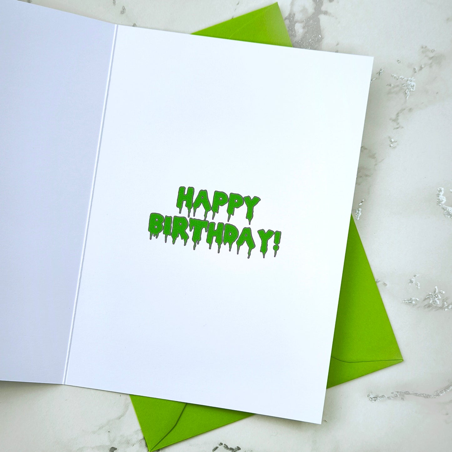 A large glossy A5 greeting card featuring a green and purple rosette with a removable 58mm badge reading "Ghoulest Mom Ever" in a bold, spooky font. Available in Mom, Mum, or Mam variations. The card is blank inside and comes with an envelope.