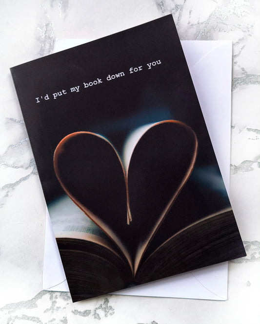 Bookish Valentine card with dark academia style. Photo of an open book with pages folded into a heart shape and a short typewritten caption reading I'd put my book down for you. Portrait format, blank inside, includes envelope.