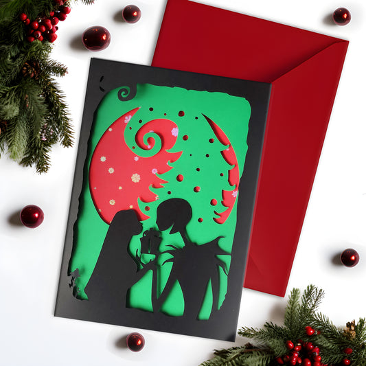 A5 handmade layered papercut Christmas card in black, red and green. Silhouette couple exchange a gift beneath a swirling tree and red moon with holographic snowflakes. Portrait format with depth. Includes red envelope. Blank inside.