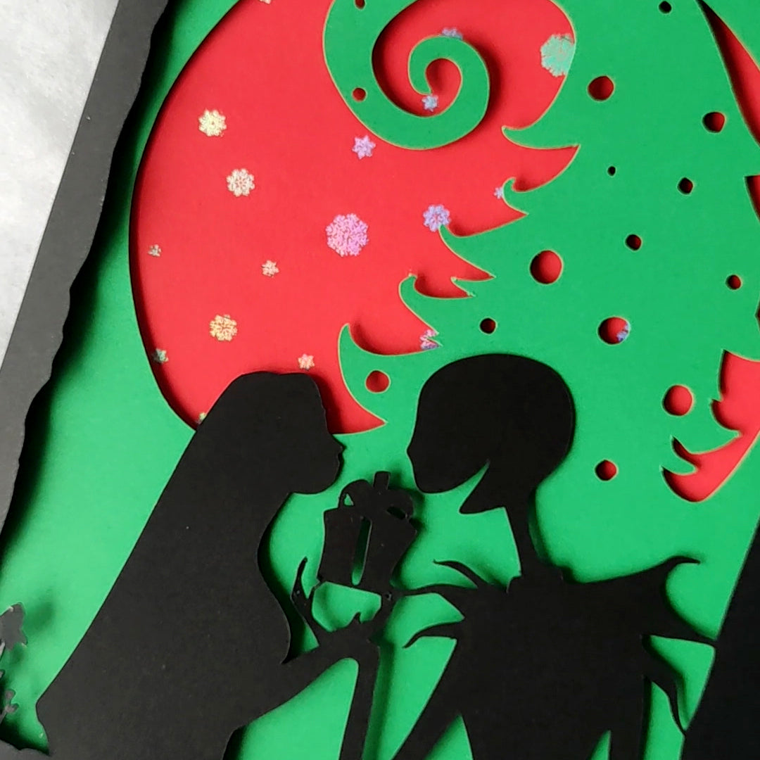 A5 handmade layered papercut Christmas card in black, red and green. Silhouette couple exchange a gift beneath a swirling tree and red moon with holographic snowflakes. Portrait format with depth. Includes red envelope. Blank inside.