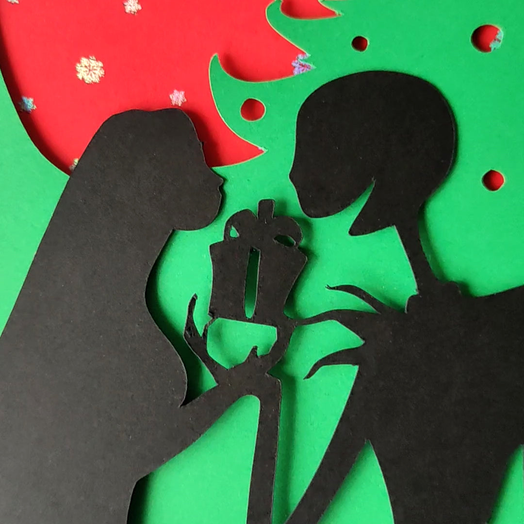 A5 handmade layered papercut Christmas card in black, red and green. Silhouette couple exchange a gift beneath a swirling tree and red moon with holographic snowflakes. Portrait format with depth. Includes red envelope. Blank inside.