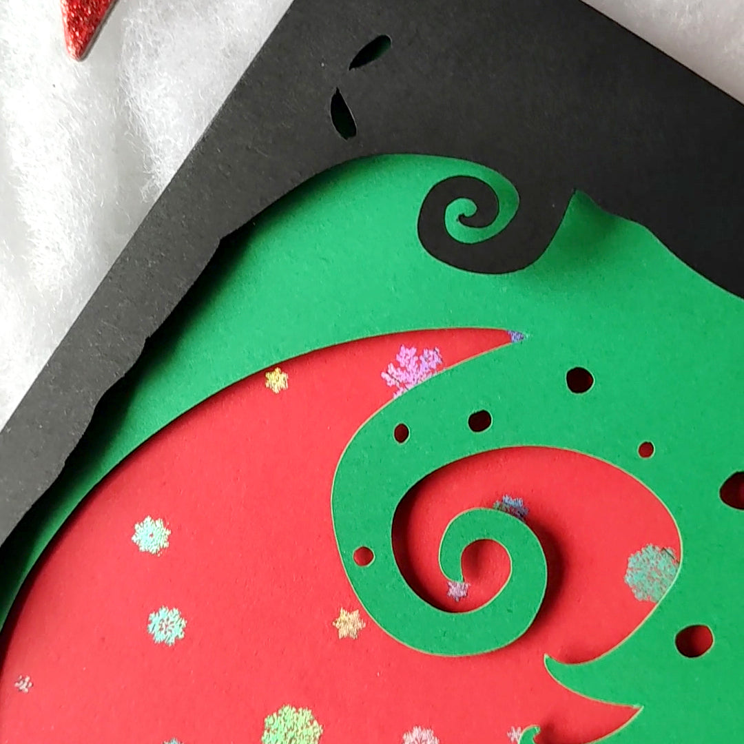 A5 handmade layered papercut Christmas card in black, red and green. Silhouette couple exchange a gift beneath a swirling tree and red moon with holographic snowflakes. Portrait format with depth. Includes red envelope. Blank inside.