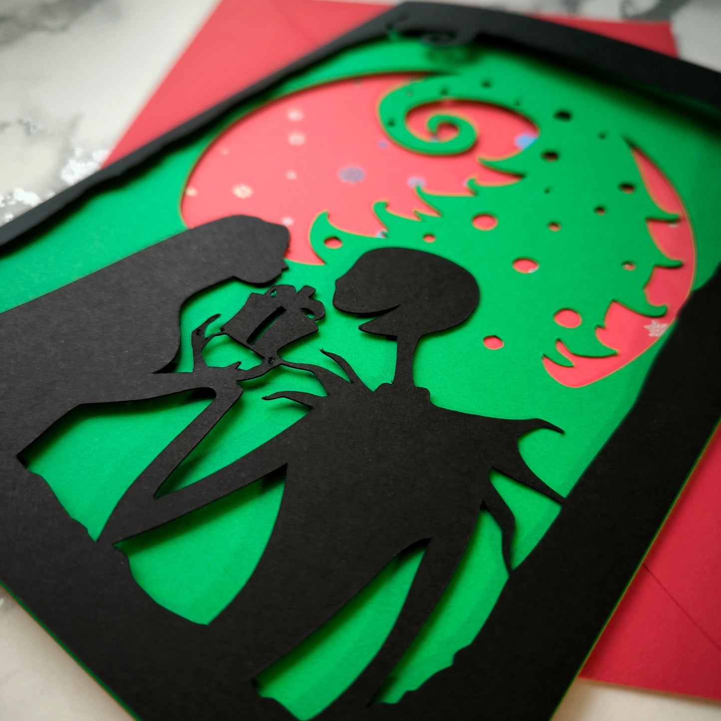 A5 handmade layered papercut Christmas card in black, red and green. Silhouette couple exchange a gift beneath a swirling tree and red moon with holographic snowflakes. Portrait format with depth. Includes red envelope. Blank inside.