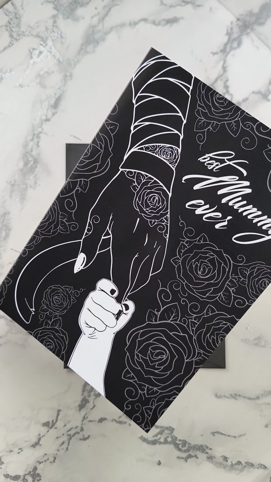 A large A5 glossy black greeting card featuring a tattoo-inspired illustration of a baby’s hand reaching for a woman’s hand wrapped in mummy bandages with rose tattoos. The card is framed by tattoo-style rose artwork. The inside is blank with matching rose motifs. Comes with an envelope included.