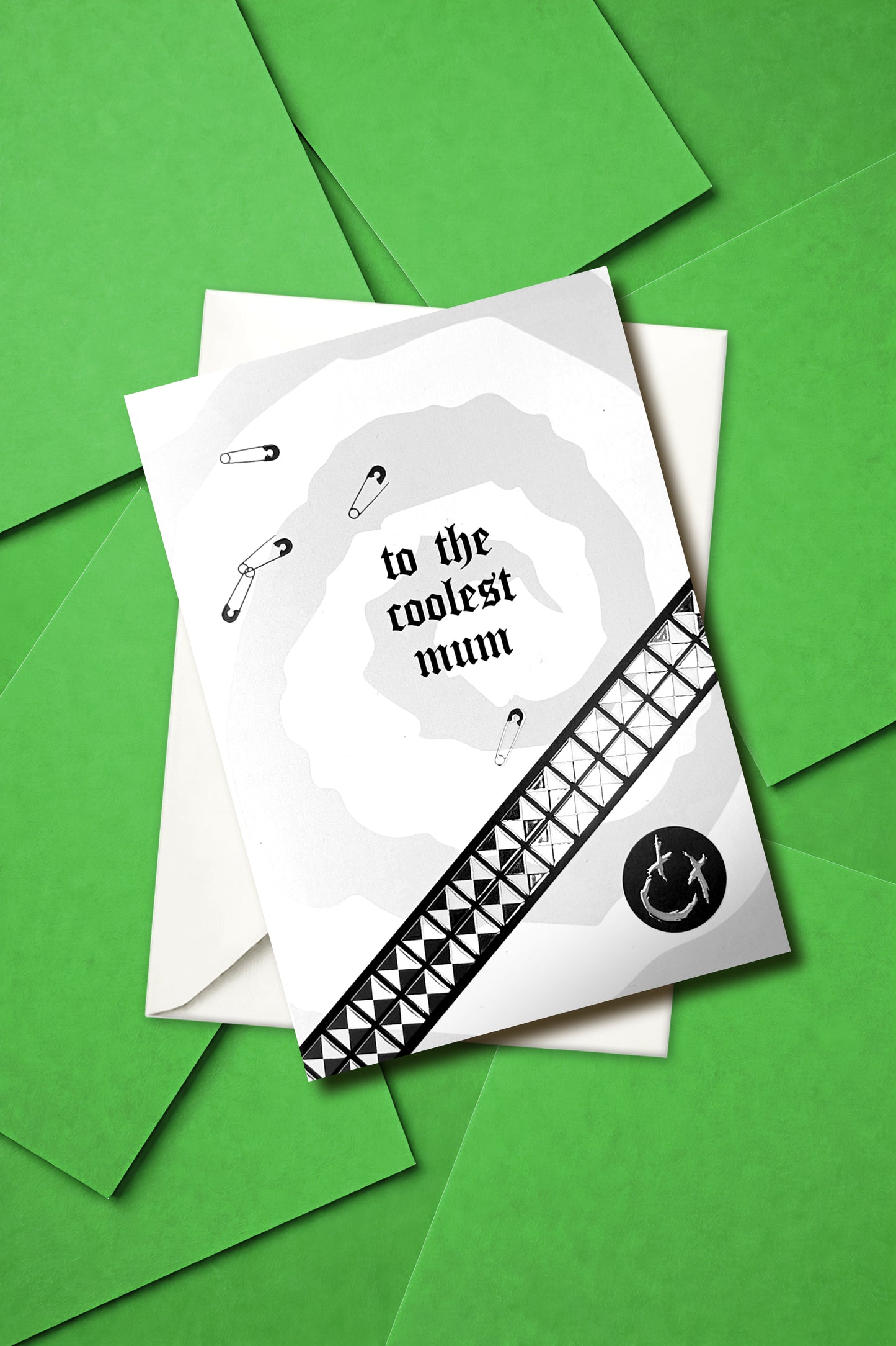 A5 greeting card reading “to the coolest mum” with gothic emo styling, featuring chrome stud belt graphic, safety pin details and a black smiley badge, paired with a white envelope.