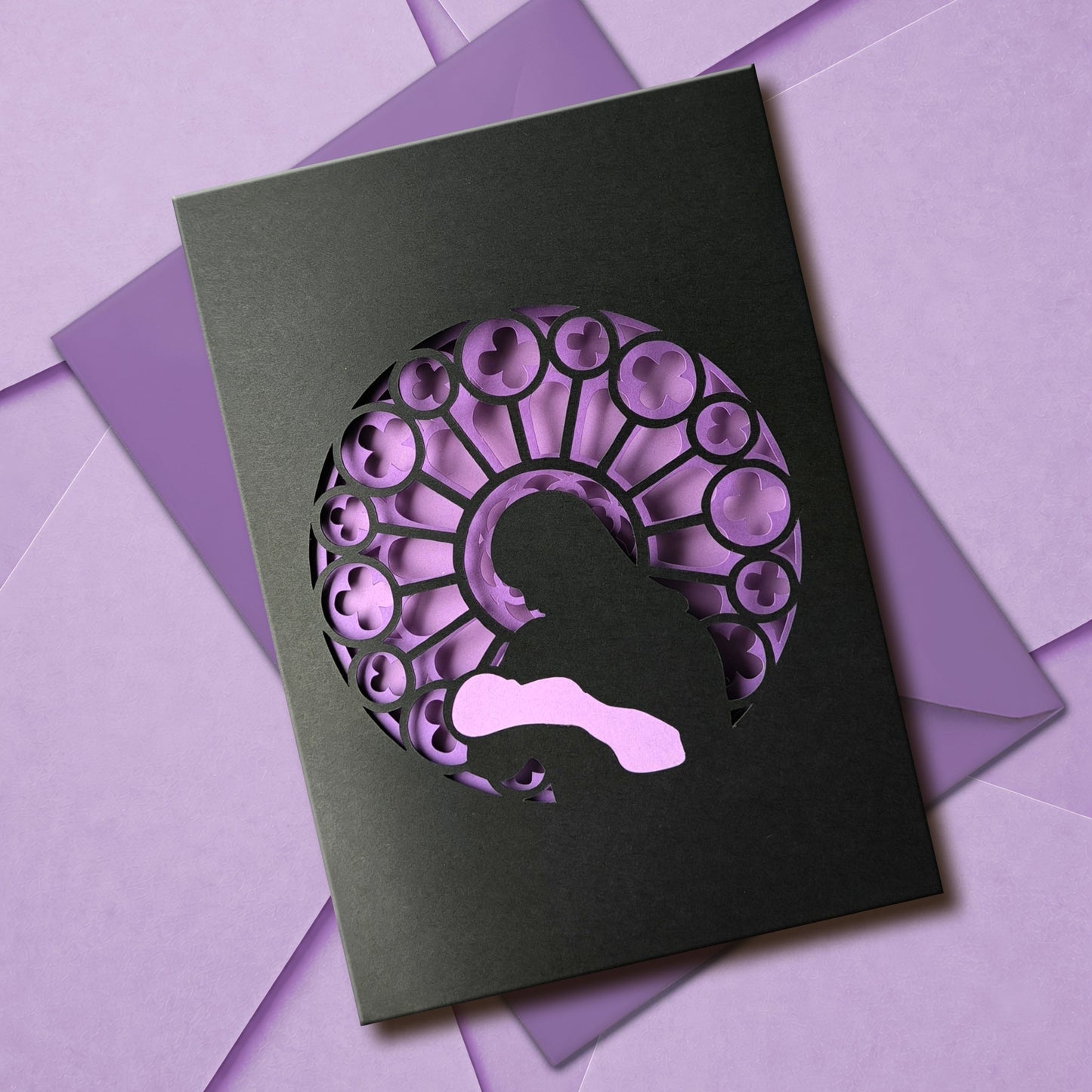 Handmade papercut greeting card, with a layered rose window in purple and black. Features a mother and baby silhouette in front of the gothic cathedral window design. Includes matching purple envelope.