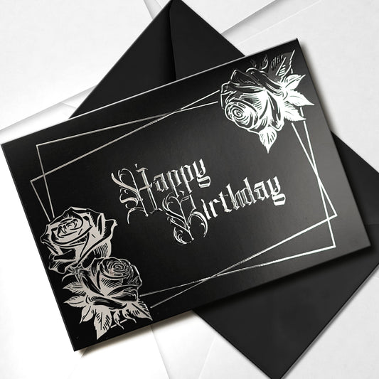 A smooth black card with raised silver chrome detailing. Silver gothic text reads Happy Birthday, surrounded by a silver rectangular frame and silver roses in two corners.