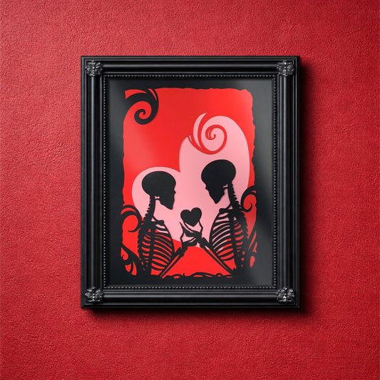 Layered papercut artwork showing two skeleton silhouettes facing each other exchanging  a small heart, set in black against red and pink heart layers with gothic flourishes. measures 8 x 10 inches. frame not included.