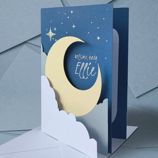 Handmade A5 layered papercut new baby card in deep navy with a cream crescent moon, white cloud layers, and silver foil star details, personalised with the phrase "welcome baby" and a custom name in silver foil script. The navy interior has a blank white cloud for your own message.