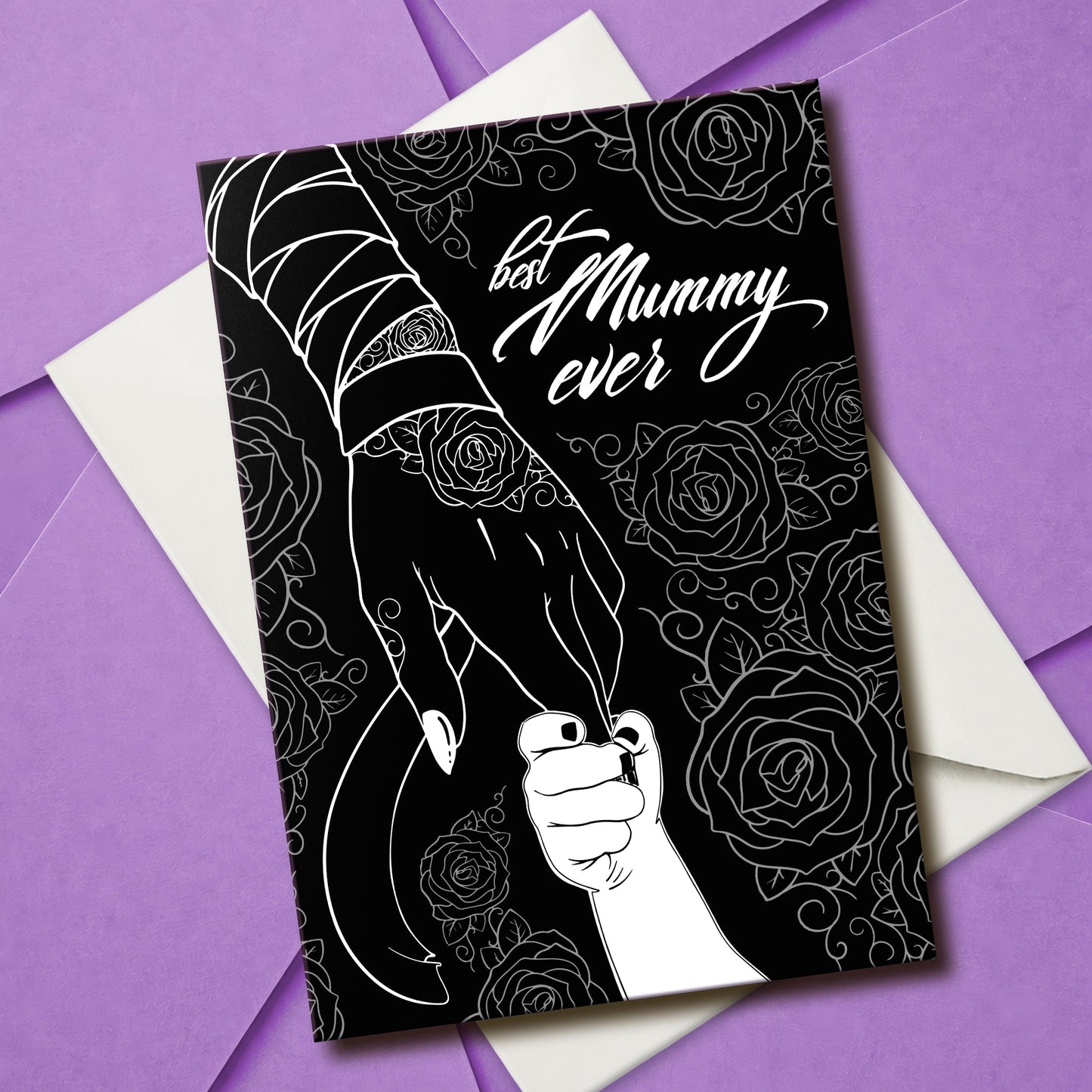 A large A5 glossy black greeting card featuring a tattoo-inspired illustration of a baby’s hand reaching for a woman’s hand wrapped in mummy bandages with rose tattoos. The card is framed by tattoo-style rose artwork. The inside is blank with matching rose motifs. Comes with an envelope included.