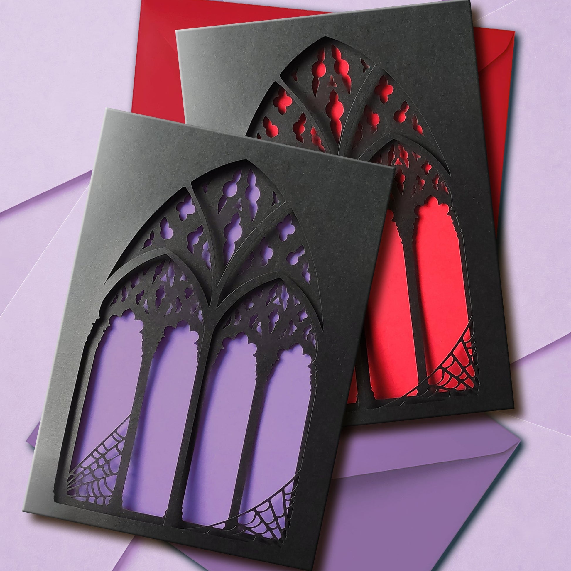 A handmade black greeting card with two intricate papercut layers of a gothic cathedral window with spiderwebs in the corners. Your choice of blood red or lilac purple shows between the window arches. Comes with a matching colour envelope.